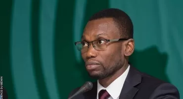 Un homme portant des lunettes et un costume sombre parle devant un micro sur fond vert