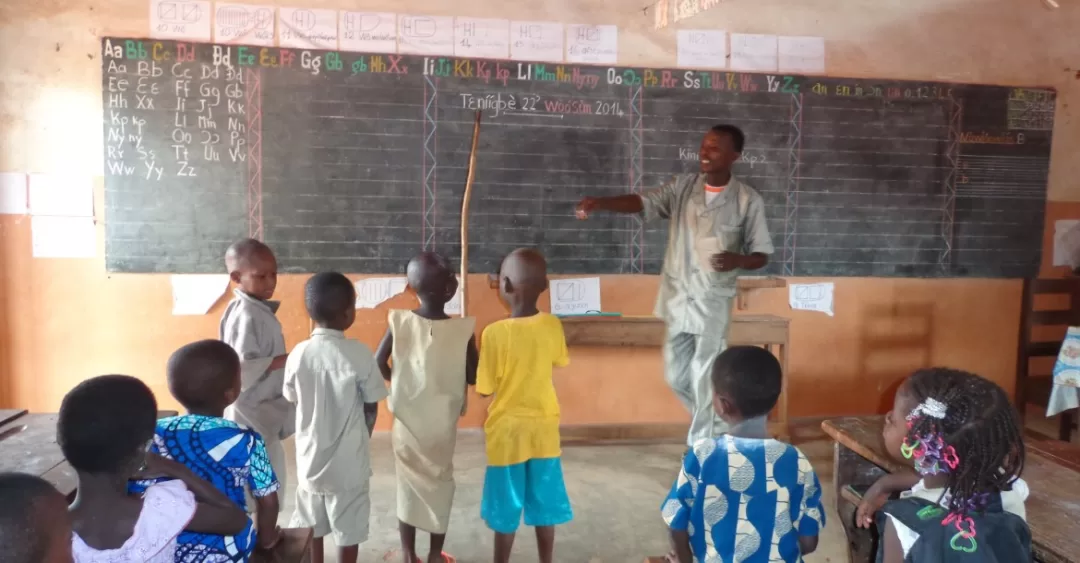 Un enseignant debout devant un tableau noir parlant à plusieurs enfants debout et assis dans une salle de classe simple