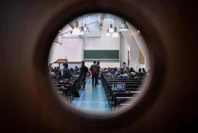 Salle de cours universitaire avec des étudiants assis et quelques-uns debout vus à travers un trou circulaire dans une porte