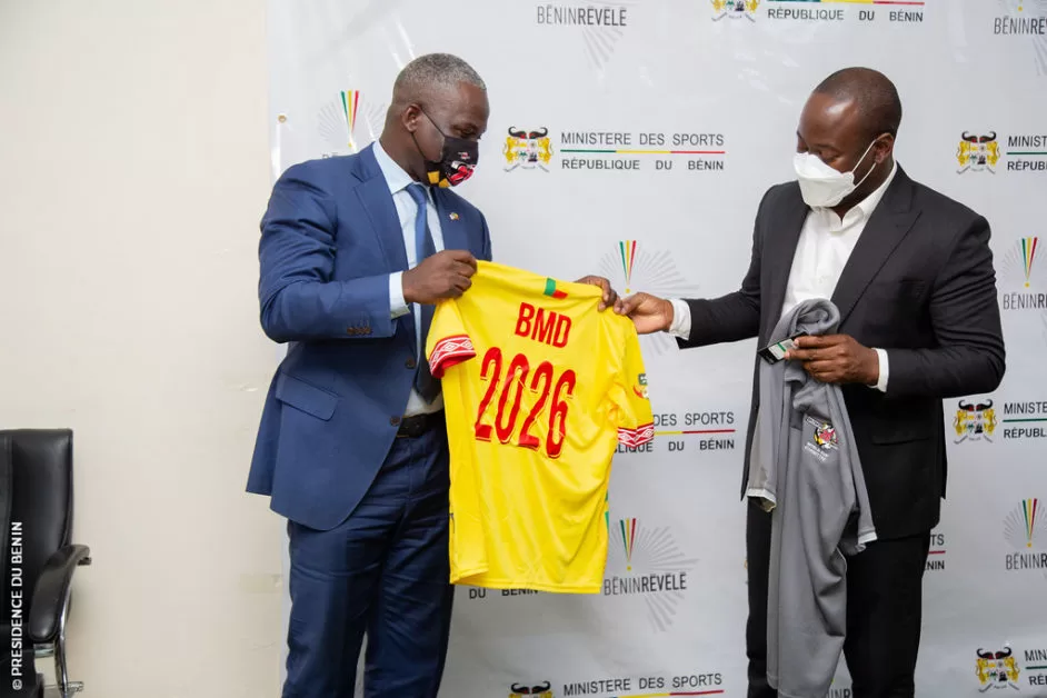 Deux hommes en costume et masque échangent des maillots devant un fond blanc marqué « Ministère des Sports République du Bénin