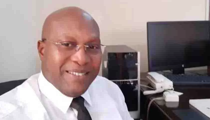 Homme portant une chemise blanche et une cravate noire souriant en bureau avec ordinateur et téléphone fixe visible en arrière-plan