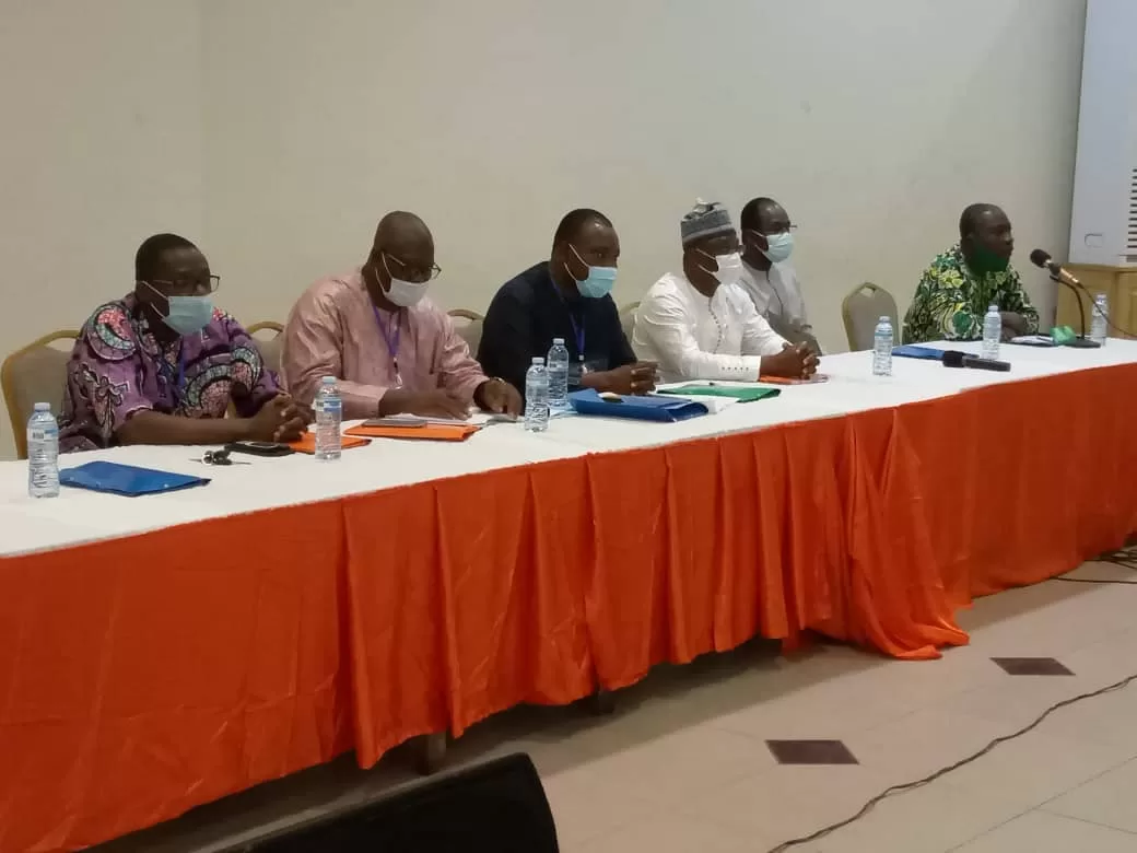 Six hommes assis à une longue table avec nappes orange et blanche portant des masques et préparant des documents
