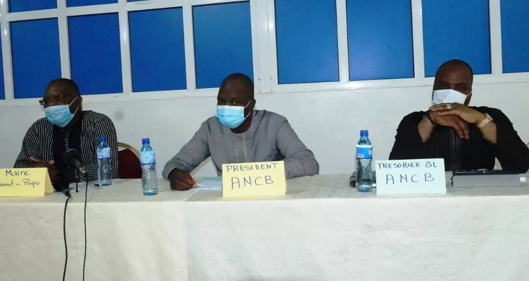 Trois hommes masqués assis à une table avec des étiquettes indiquant leurs fonctions dans une réunion formelle