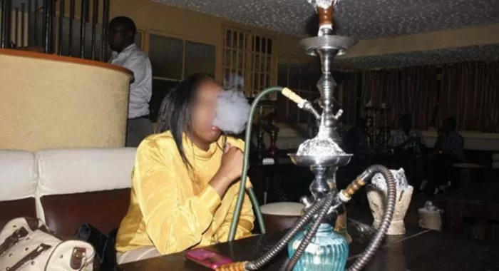 Une personne fumant une chicha dans un intérieur sombre avec plusieurs narguilés posés sur une table devant elle