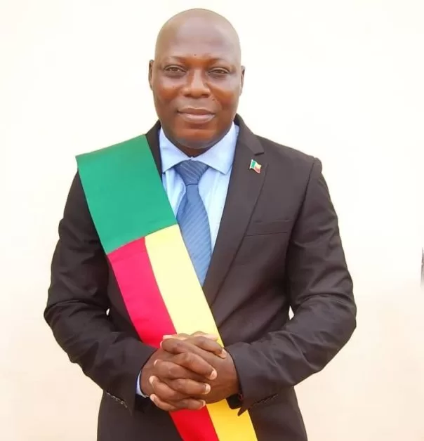 Homme en costume sombre avec une écharpe tricolore vert, jaune et rouge posant devant un fond blanc