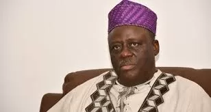 Homme en tenue traditionnelle africaine et chapeau violet, assis sur un fauteuil marron claire