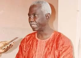 Homme âgé en tenue traditionnelle rouge parlant face à un microphone tenu par une main extérieure