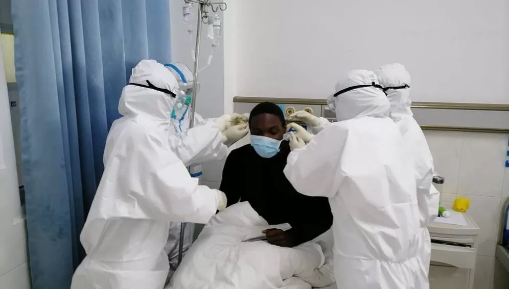 Deux soignants en tenue de protection complète entourent un patient masqué allongé dans un lit d'hôpital
