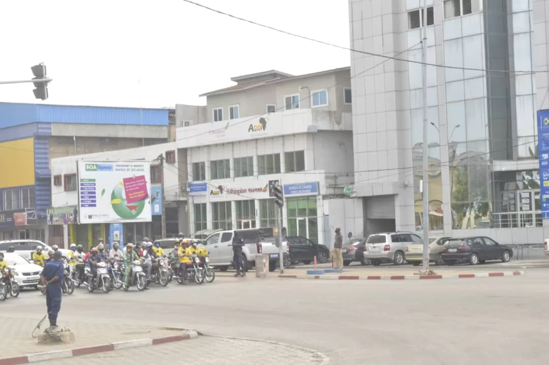 Un groupe de motards et plusieurs piétons se tiennent à un feu tricolore dans un carrefour urbain devant des bâtiments modernes
