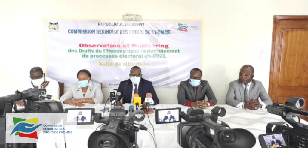 Cinq personnes masquées assises derrière une table face à plusieurs caméras dans une salle de conférence avec une bannière en arrière-plan.