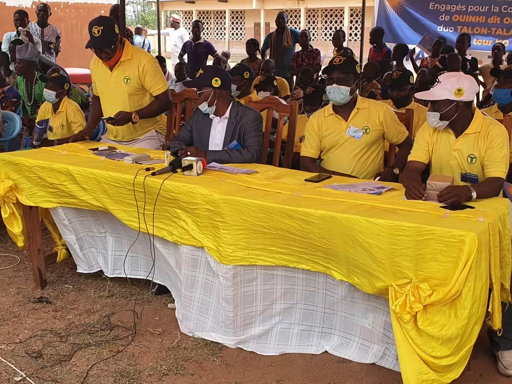 Personnes assises à une table couverte de tissu jaune et blanc, portant des polos jaunes et casquettes noires avec logo, en extérieur sous abri