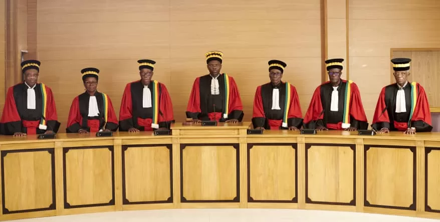 Sept membres en tenue officielle rouge et noire debout derrière un bureau en bois clair dans une salle officielle