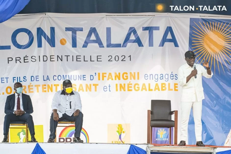Trois hommes sur scène dont un debout parlant devant une grande banderole électorale aux couleurs bleu, blanc et orange