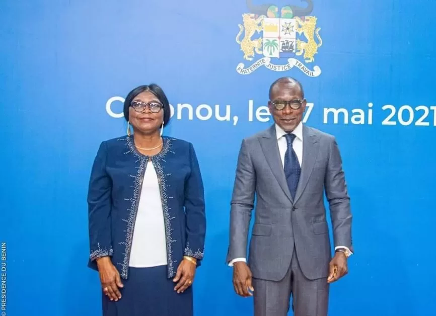 Une femme en tailleur bleu et un homme en costume gris posent devant un mur bleu portant un blason et un texte dates