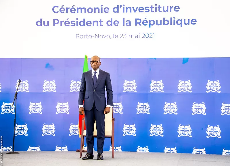 Un homme en costume sombre se tient debout devant un fond bleu avec texte et armoiries blanches et un drapeau visible derrière lui
