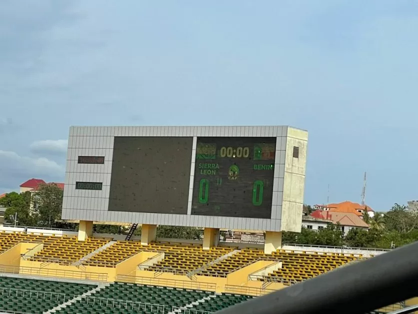 Un tableau d'affichage dans un stade indique 0-0 et le chronomètre à 00:00 au-dessus des gradins jaunes et verts