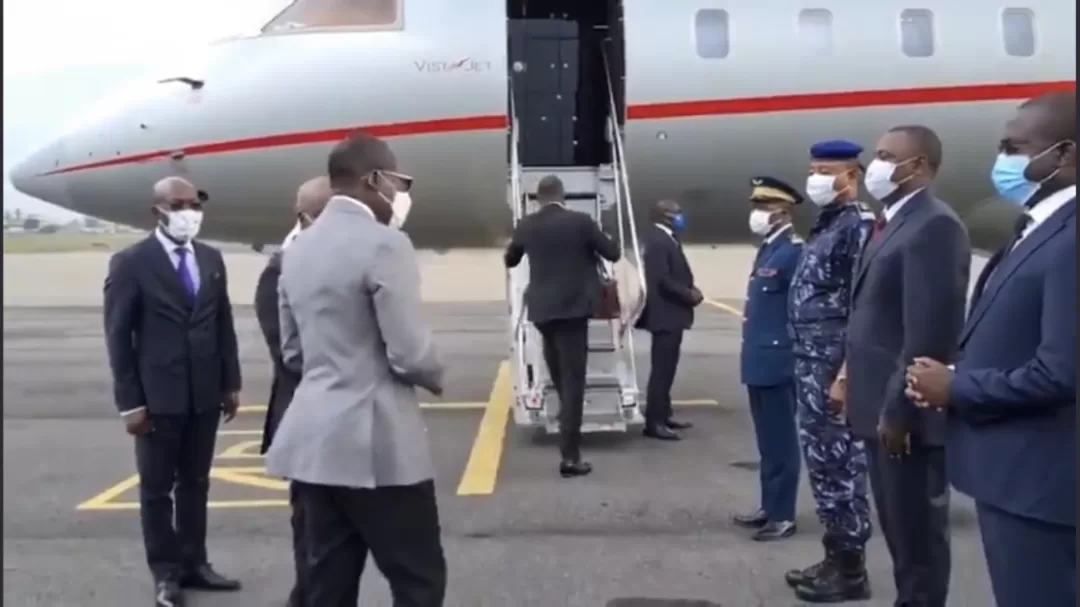 Homme en costume montant les marches d’un avion privé entouré de plusieurs personnes en costume et uniforme portant un masque