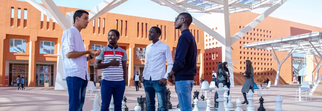 Quatre jeunes hommes discutent autour d’un jeu d’échecs géant en plein air dans une cour moderne ensoleillée