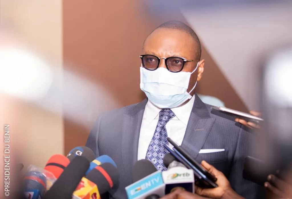 Homme en costume, lunettes et masque blanc parlant devant plusieurs micros de médias