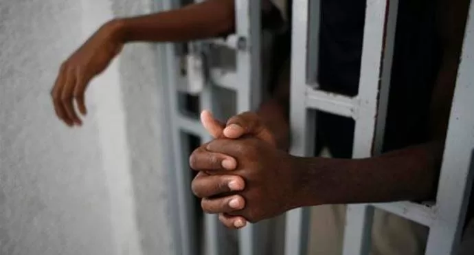 Mains croisées d’une personne derrière des barreaux avec un bras posé sur la barre métallique verticale