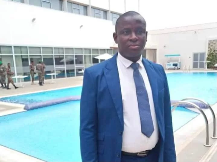 Un homme en costume bleu posé debout devant une piscine avec un bâtiment en arrière-plan et des militaires à distance