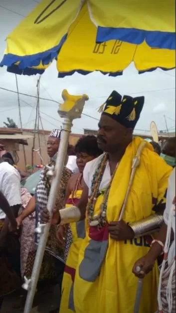Un homme en habits traditionnels jaunes et or tenant un sceptre sous un grand parasol jaune entouré de plusieurs personnes