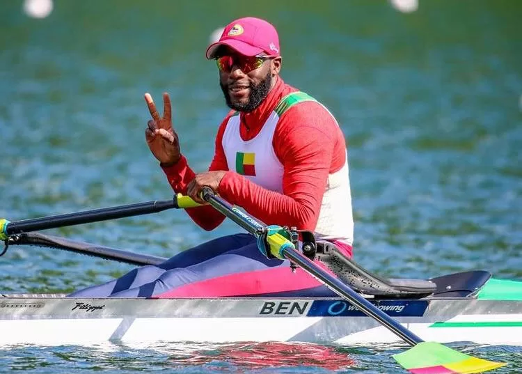 Rameur en tenue sportive rouge et blanche avec drapeau sur un bateau d’aviron faisant un signe de victoire sur l’eau