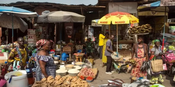 Marché animé avec des vendeuses et acheteurs autour de divers produits alimentaires en plein air sous parasols.