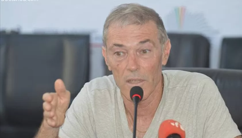 Homme en chemise claire parlant devant un micro lors d’une conférence avec un fond flou et un autre micro rouge devant lui