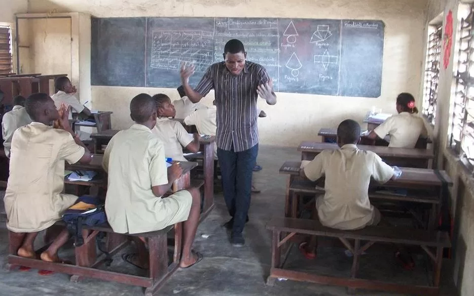 Un enseignant debout face à une classe d’élèves assis sur des bancs dans une salle avec un tableau noir écrit derrière lui