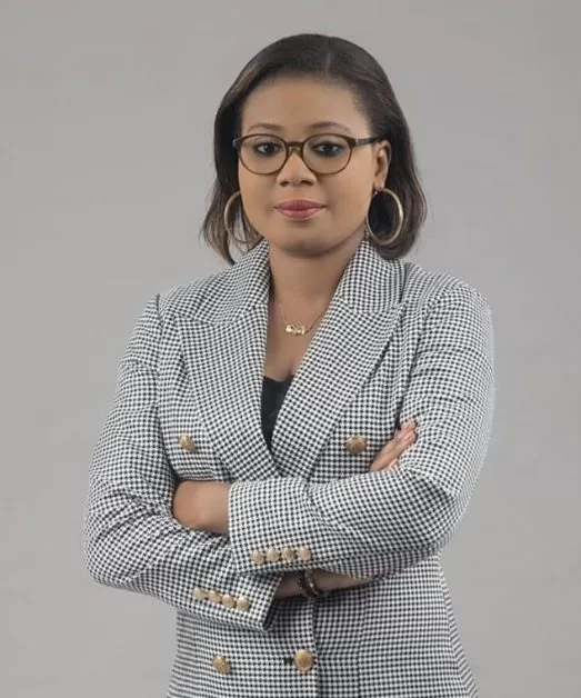 Femme portant un blazer à carreaux et des lunettes, bras croisés, regardant vers l'avant sur fond gris