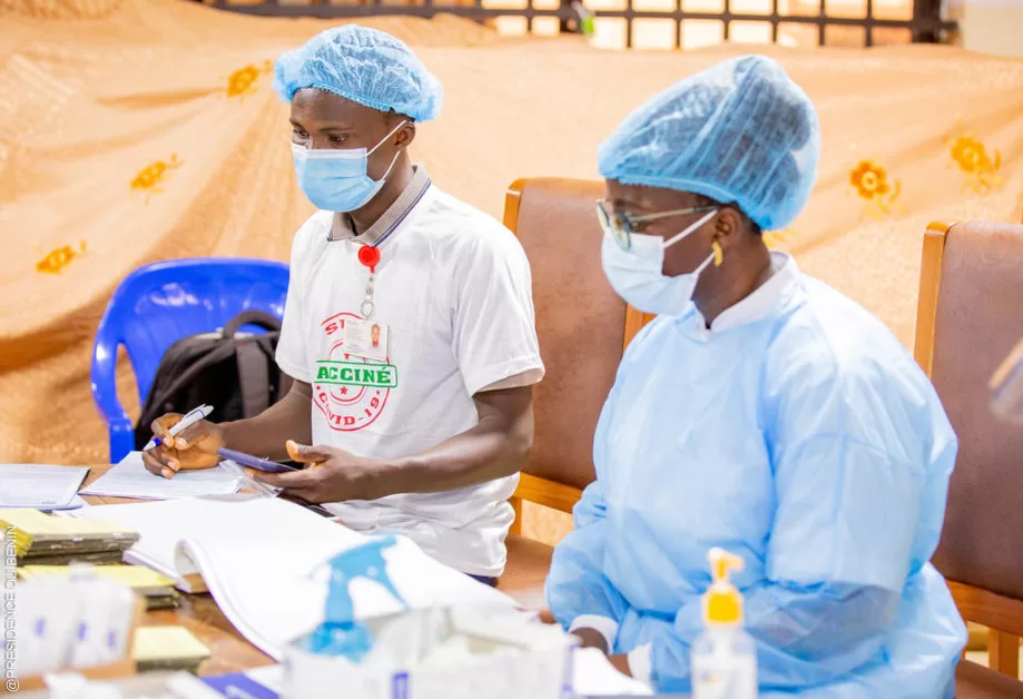 Deux professionnels de santé portant blouse, charlotte et masque sont assis devant un bureau avec documents et flacons sanitaires.