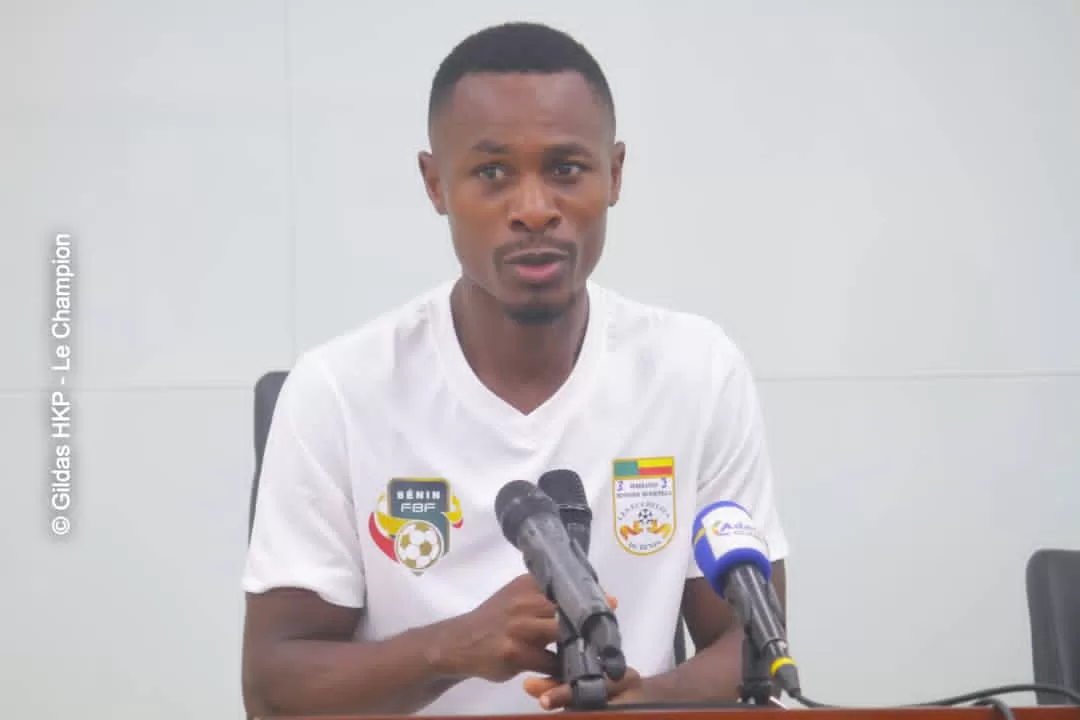 Un homme en polo blanc avec logos de football parle devant deux micros lors d'une conférence de presse intérieure