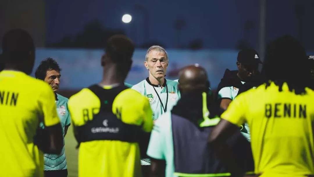 Un homme au centre parle à un groupe de joueurs en maillot jaune marqué BENIN lors d’un entraînement en soirée