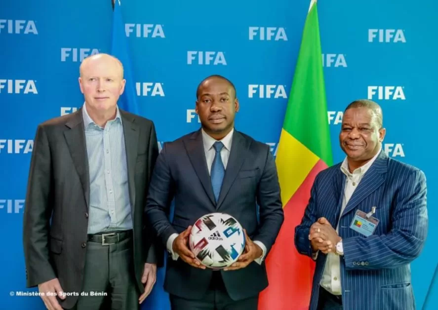 Trois hommes en costume posent devant un drapeau tricolore avec un ballon de football devant un fond bleu marqué FIFA
