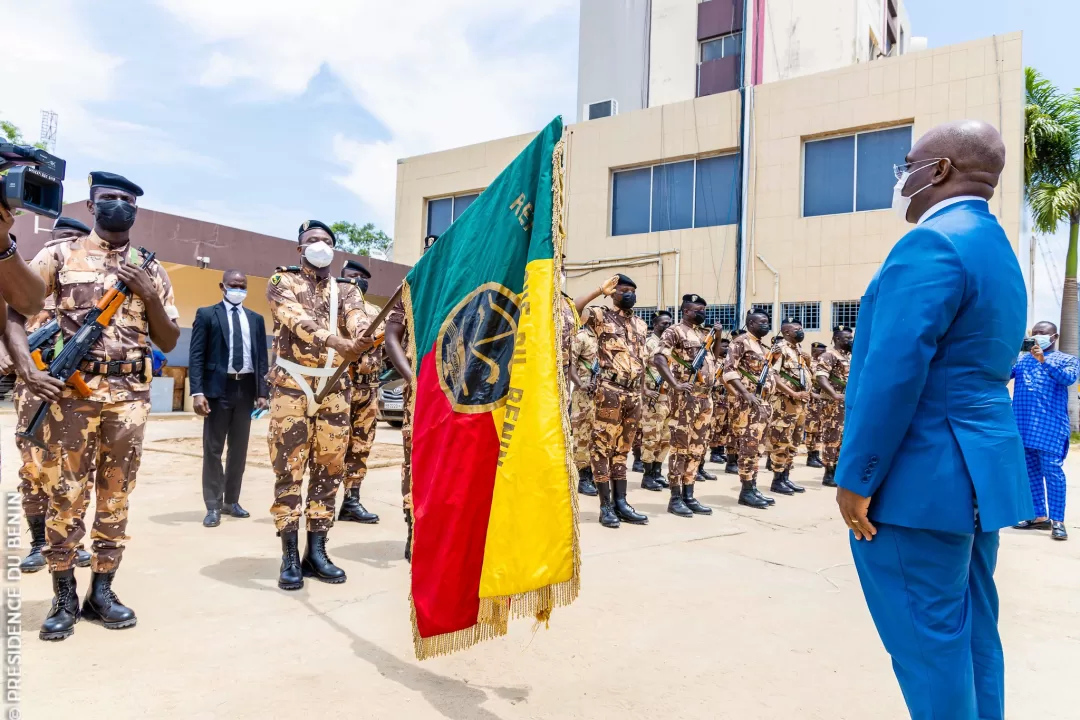 Des soldats en uniforme camouflage, certains armés, en formation devant un homme en costume bleu et masque blanc devant un bâtiment moderne