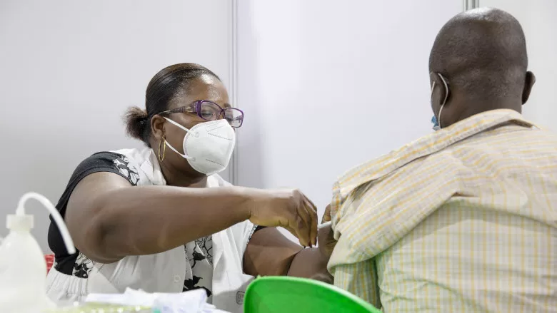 Une femme en blouse et masque vaccine un homme vu de dos assis sur une chaise verte dans un centre de vaccination