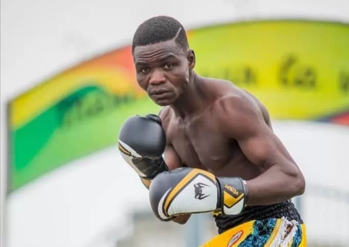 Boxeur torse nu en garde de combat avec gants noirs et jaunes sur un ring en extérieur coloré en arrière-plan