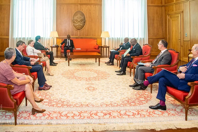 Plusieurs personnes assises en cercle dans une grande salle avec tapis rouge et beige et murs en bois clair
