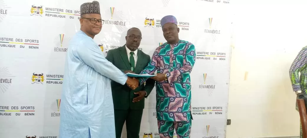 Trois hommes en costume traditionnel et costume formel se serrent la main devant un mur avec logos officiels liés au sport du Bénin