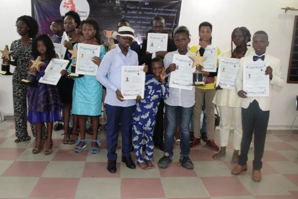 Un groupe d’enfants posant debout, tenant des certificats et des trophées en forme d’étoile dans une salle.
