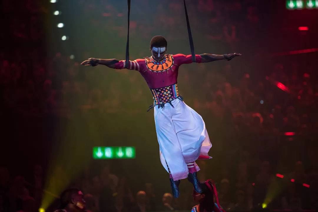 Un artiste vêtu d’un costume coloré et maquillé, suspendu dans les airs lors d’un spectacle devant un public nombreux