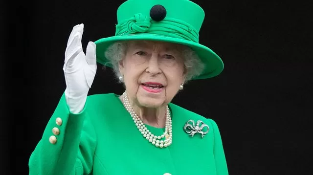 Personne âgée souriante en manteau vert avec chapeau assorti, gant blanc, collier de perles et broche, saluant de la main droite face caméra