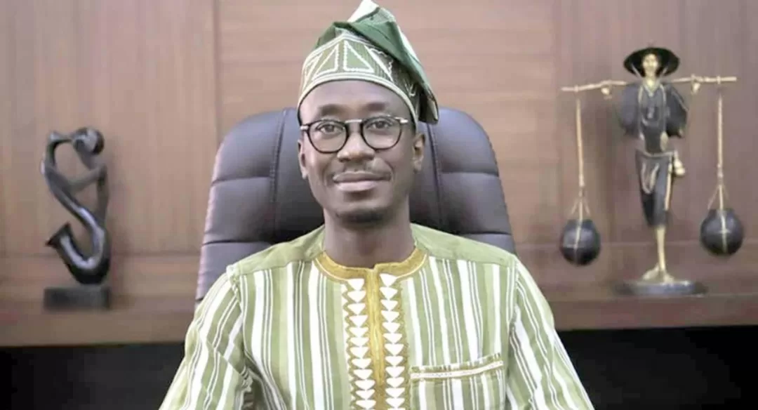 Homme portant une tenue traditionnelle rayée verte et blanche avec un chapeau assorti assis devant un bureau avec des sculptures en arrière-plan