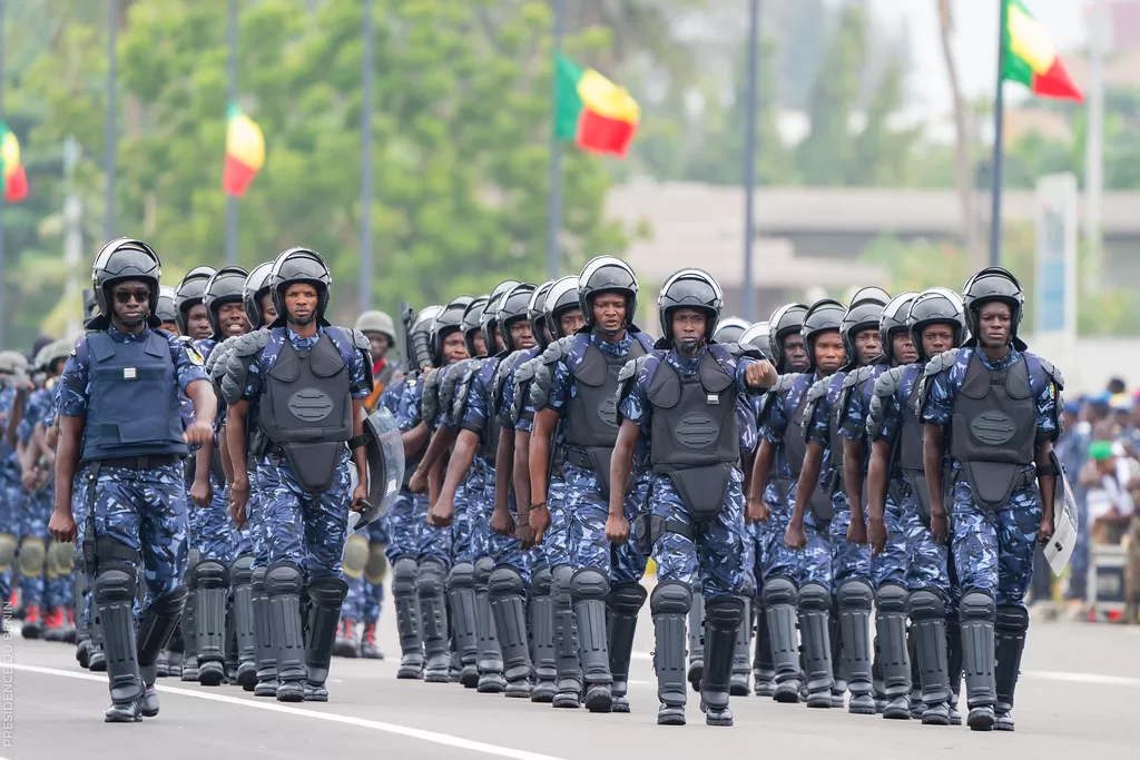 Des agents en tenue tactique et casque défilent en formation serrée sur une avenue bordée de drapeaux colorés