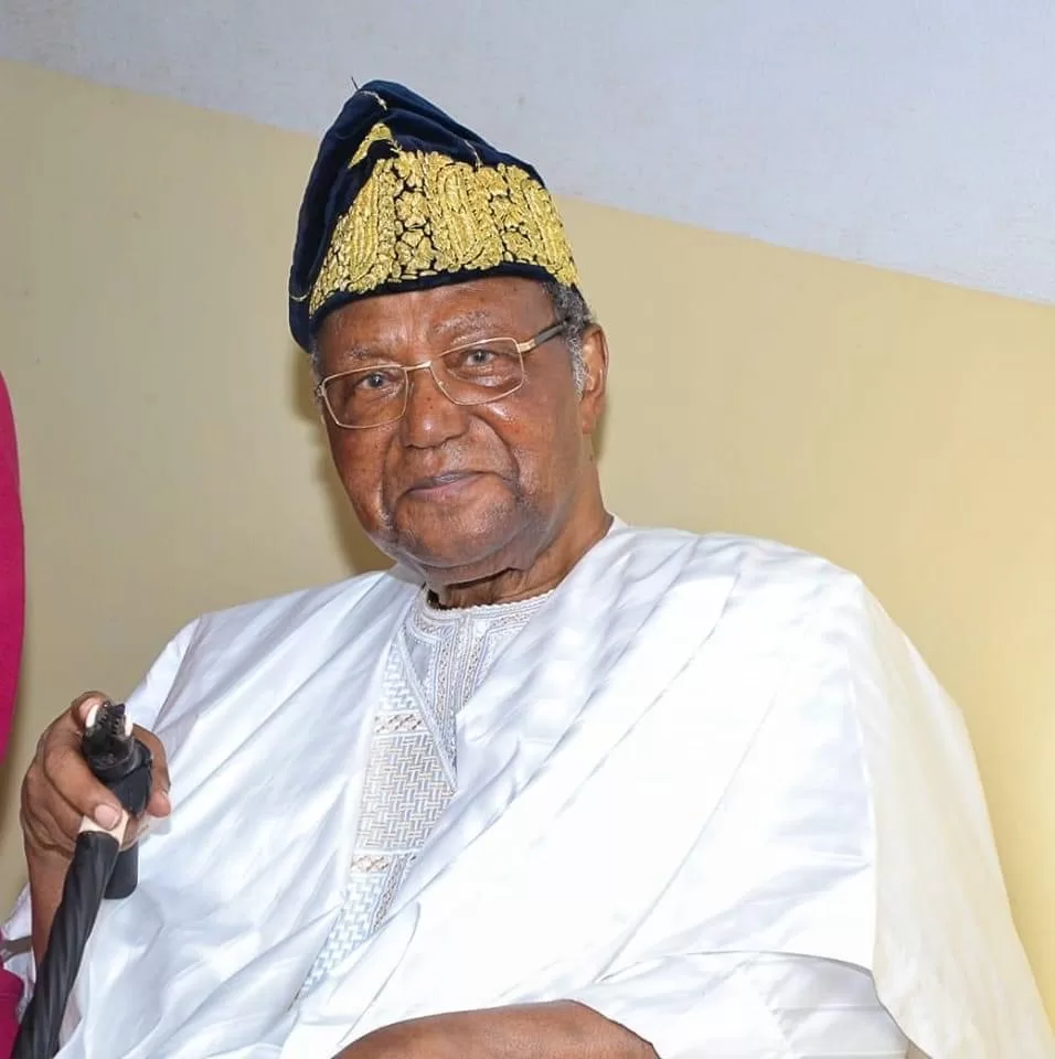 Homme âgé portant une tenue traditionnelle blanche et un chapeau brodé tenant une canne assis devant un mur clair