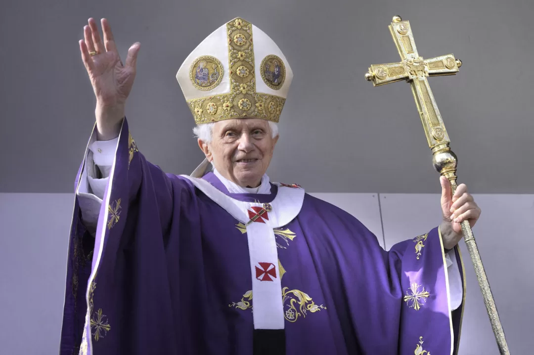 Un homme âgé en tenue ecclésiastique pourpre avec une mitre dorée lève la main droite et tient une grande croix dorée de la main gauche