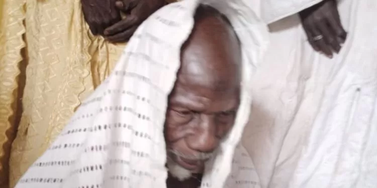 Un homme âgé en tenue traditionnelle blanche avec un tissu sur la tête entouré d'autres personnes en habits clairs.