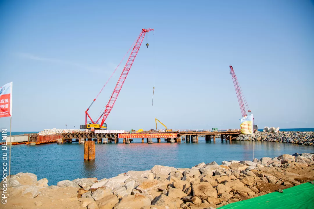 Deux grues rouges installées sur une structure en construction au-dessus de l'eau avec des rochers au premier plan