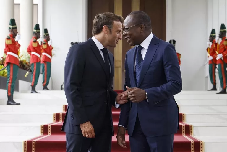 Deux hommes en costume échangent en souriant devant un escalier rouge orné de gardes en uniforme vert et rouge.